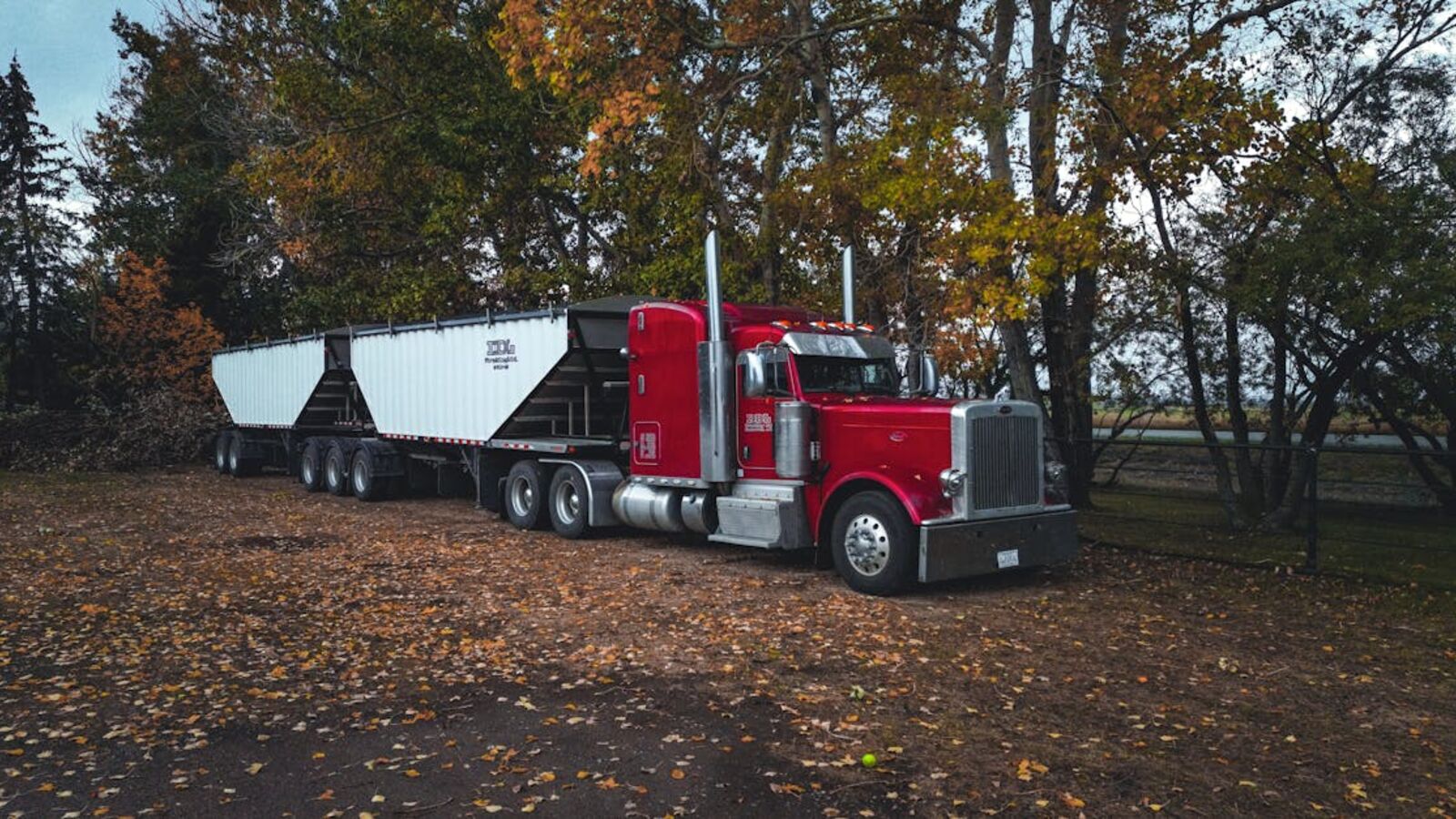 Выкуп тягачей в Санкт-Петербурге — A red trailer truck parked outdoors surrounded by autumn trees in Purple Springs, Canada.