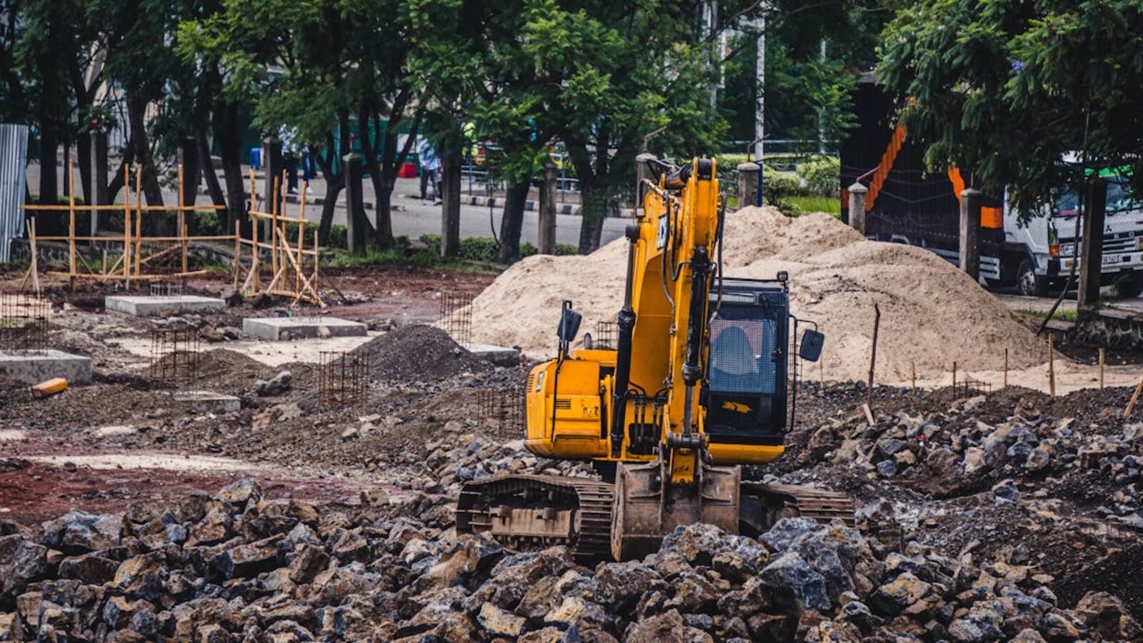 A yellow excavator working at an urban construction site surrounded by rocks and trees.